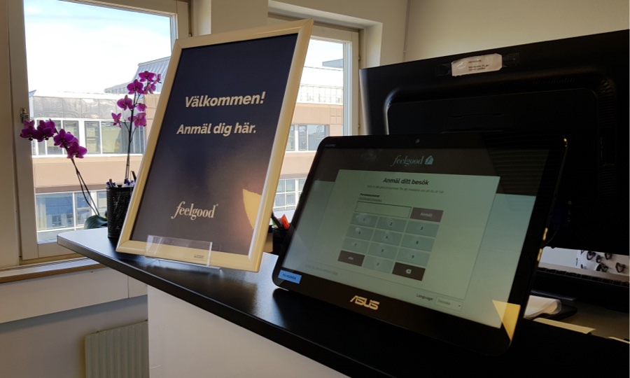 The tablet running Feelgood Visitor Registration standing on a reception desk next to a poster telling visitors to register here.